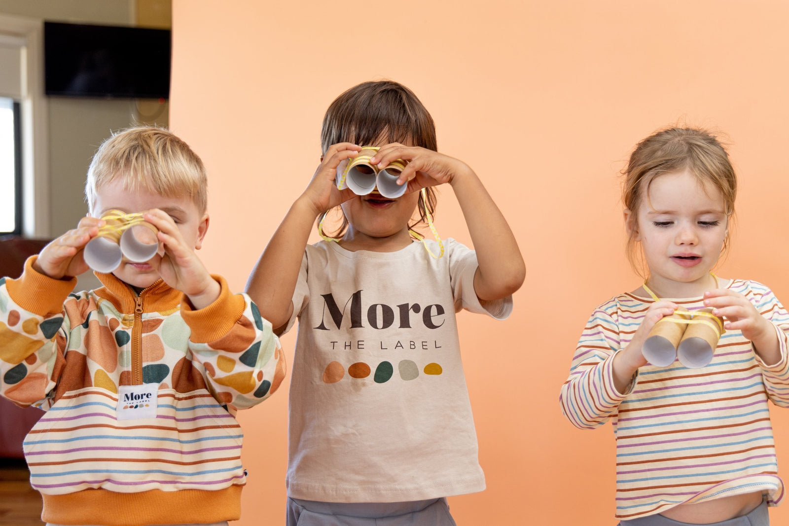 More the label hero image of three children looking through home made binoculars. Boy on the right wears More the label stone print pullover. Boy in the middle wears logo tee with dusty blue track pants and girl to the right wears striped long sleeve tee with dusty blue track pants.