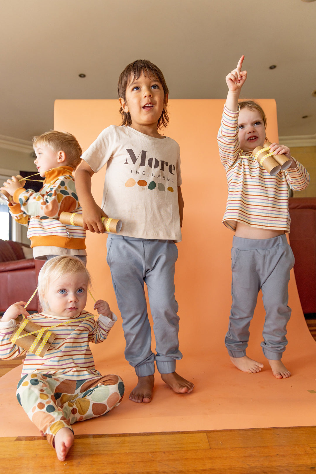 Four children standing on photography set with homemade binoculars. First child wears More the label stone print pullover, logo tee and dusty blue track pant. Second child wears dusty blue track pant and logo tee. Third child wears dusty blue track pant and striped long sleeve tee and fourth child wears stone print leggings and striped long sleeve tee.  
