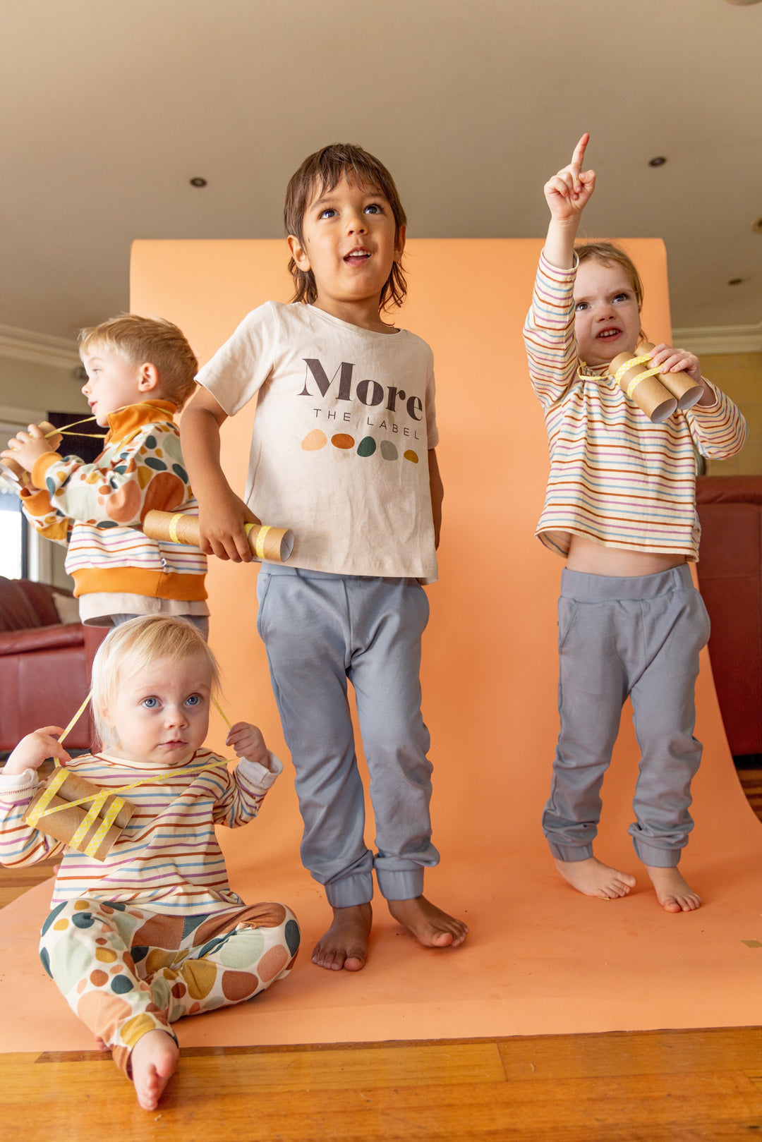 Four children standing on photography set with homemade binoculars. First child wears More the label stone print pullover, logo tee and dusty blue track pant. Second child wears dusty blue track pant and logo tee. Third child wears dusty blue track pant and striped long sleeve tee and fourth child wears stone print leggings and striped long sleeve tee.