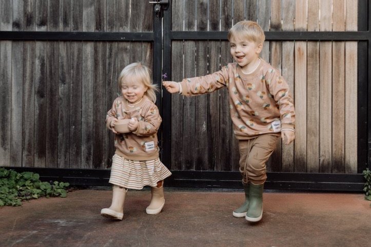 Image of two children running and laughing wearing More the label. First child is wearing stone huddle jumper over the top of the striped skater dress with gumboots. Second child is wearing stone huddle jumper and chocolate brown track pants.