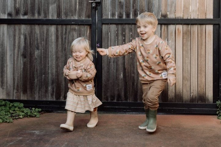 Photo of two children laughing and running. First child wears More the label striped skater dress with stone print leggings and stone huddle jumper. Second child wears chocolate track pants with stone huddle jumper.  