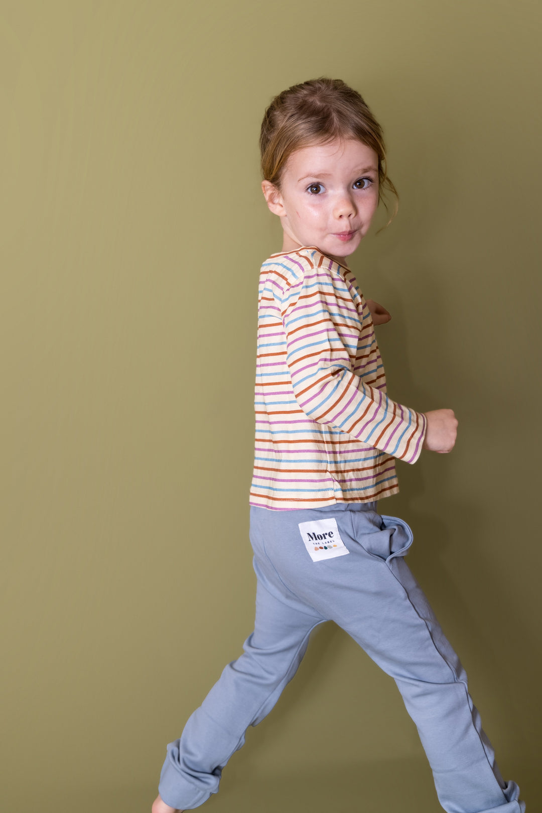 Full length image of three year old girl with cheeky look on her face wearing More the label striped long sleeve tee and dusty blue track pant. 