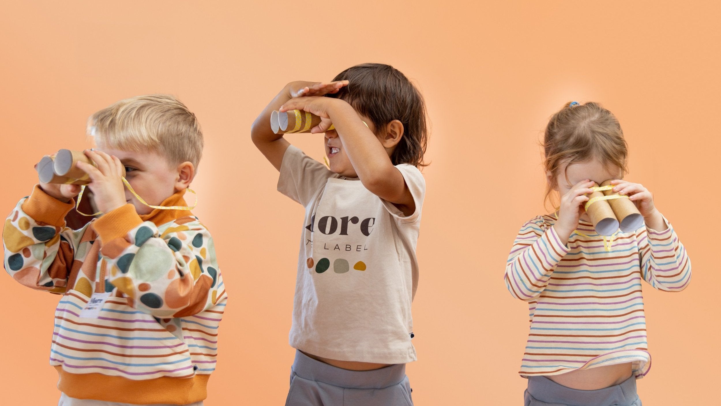 More the label hero image of three children looking through home made binoculars. Boy on the right wears More the label stone print pullover. Boy in the middle wears logo tee with dusty blue track pants and girl to the right wears striped long sleeve tee with dusty blue track pants. 