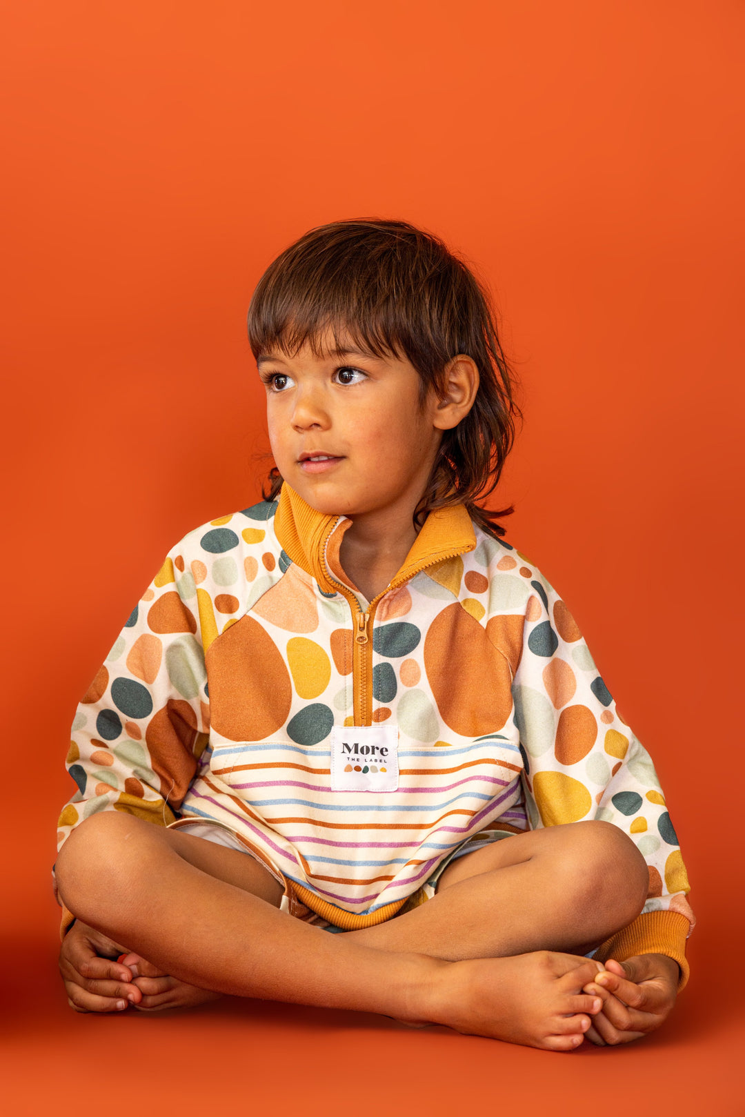 Photo of four year old boy sitting cross legged and looking to the side on a red background. Boy wears More the label stone print pullover and stone print bike short. 