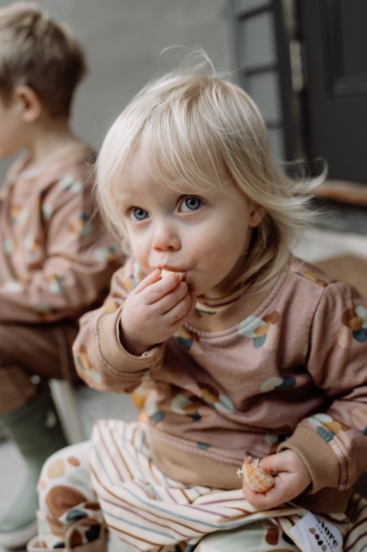 Image of child eating mandarine while wearing More the label stone huddle jumper over the striped skater dress with stone print leggings underneath. 