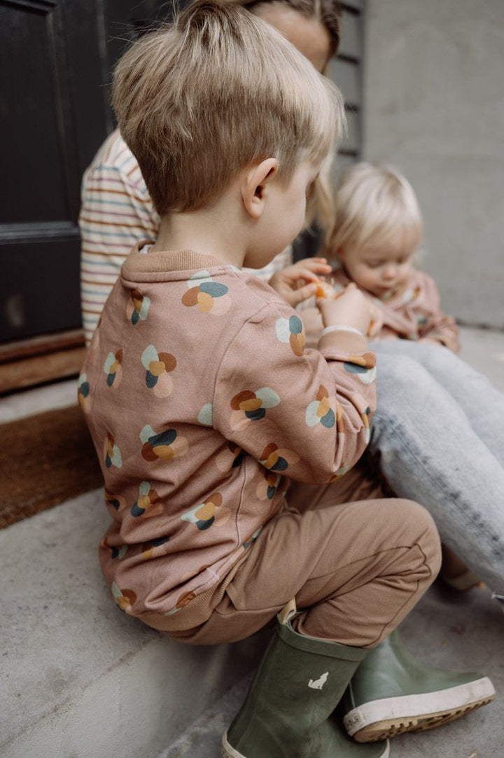Photo of children and mother sitting on a step eating mandarins. First child wears More the label stone huddle jumper with chocolate track pant. Second child is wearing stone huddle jumper and mother is wearing striped long sleeve top. 