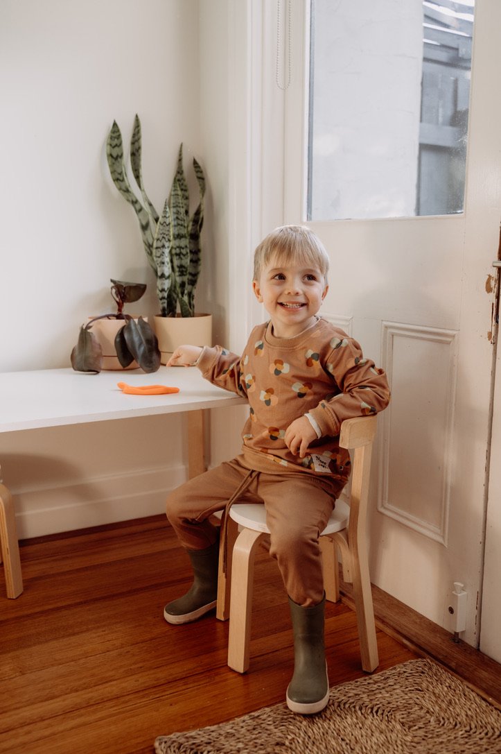 Photo of four year old boy sitting on white chair at white table wearing More the label chocolate track pant with stone huddle jumper.