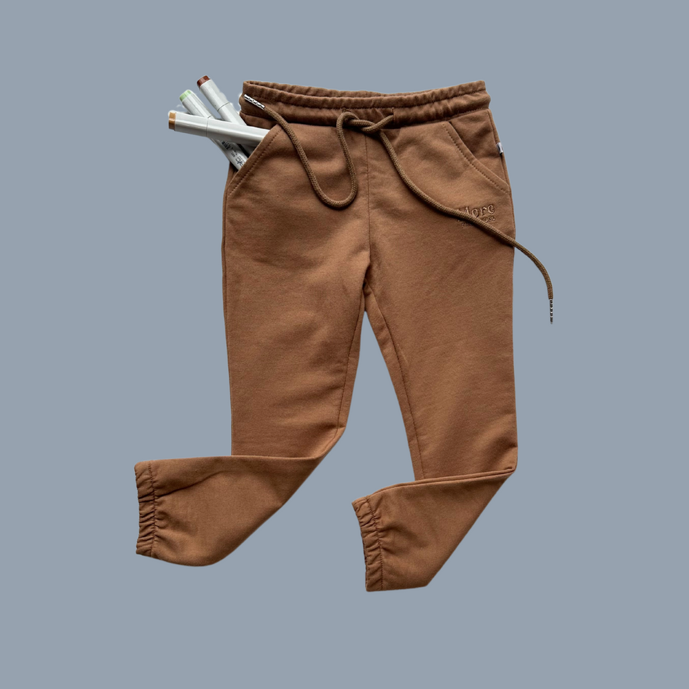 More The Label organic cotton children's clothing chocolate track pant flat lay image of front with legs flicked to the side and textas in pocket on on a blue background. Brown track pants with elastic waste and drawstring. 