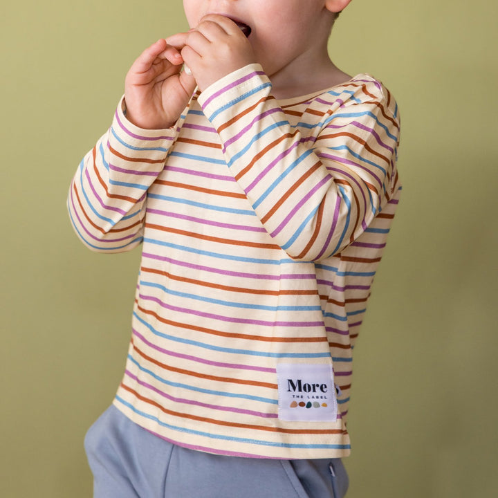 Close up image of child wearing More the label striped long sleeve tee with dusty blue track pants. Hands in mouth eating. 