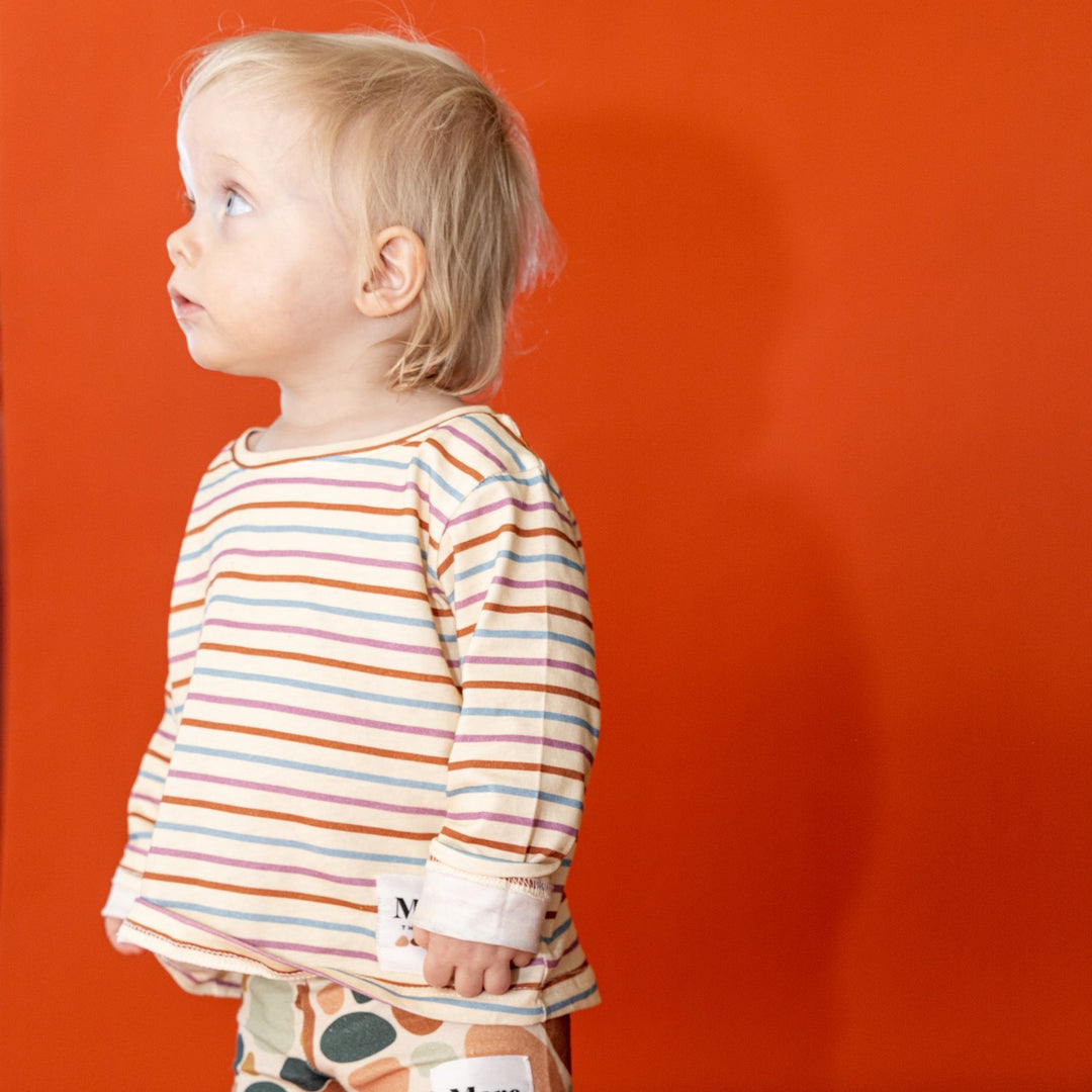Image of one year old child in front of red backdrop looking side on wearing More the label striped long sleeve tee and stone print bike shorts. 