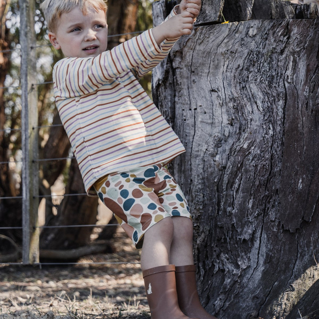 Photo of three year old child handing off old tree stump in the bush wearing More the label wearing stone print bike shorts with striped long sleeve tee and cry wolf gumboots